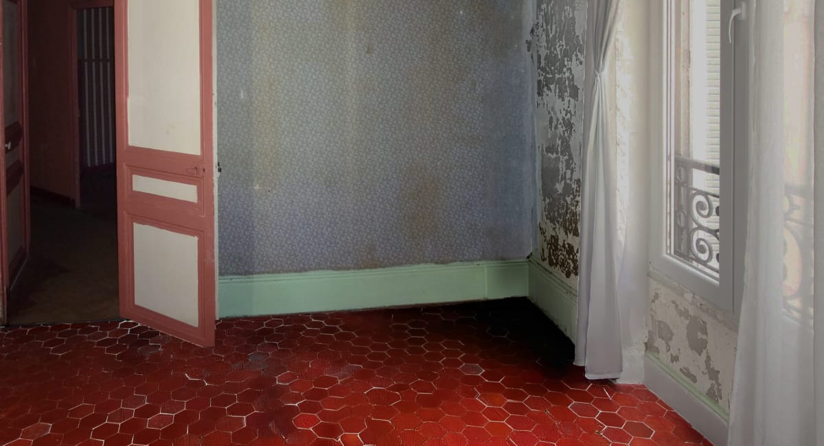 Une pièce délabrée avec un papier peint décollé, un sol en carreaux rouges et une fenêtre ancienne.
