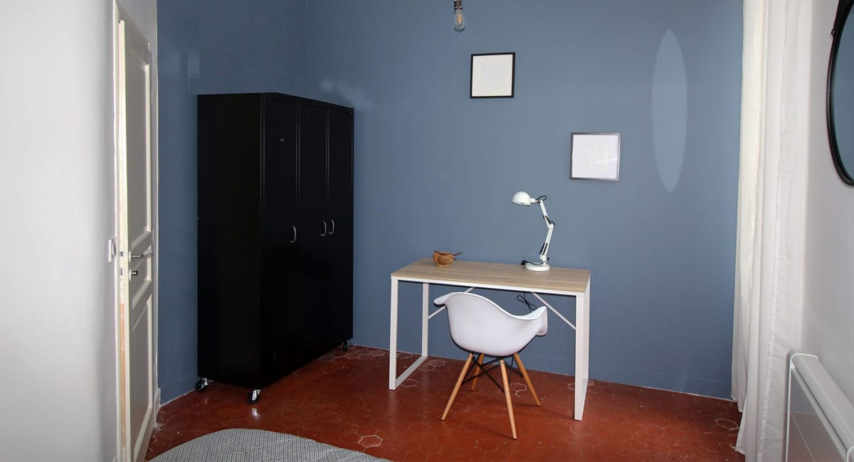 Une petite pièce avec un bureau, une chaise et un placard noir sur un mur bleu.