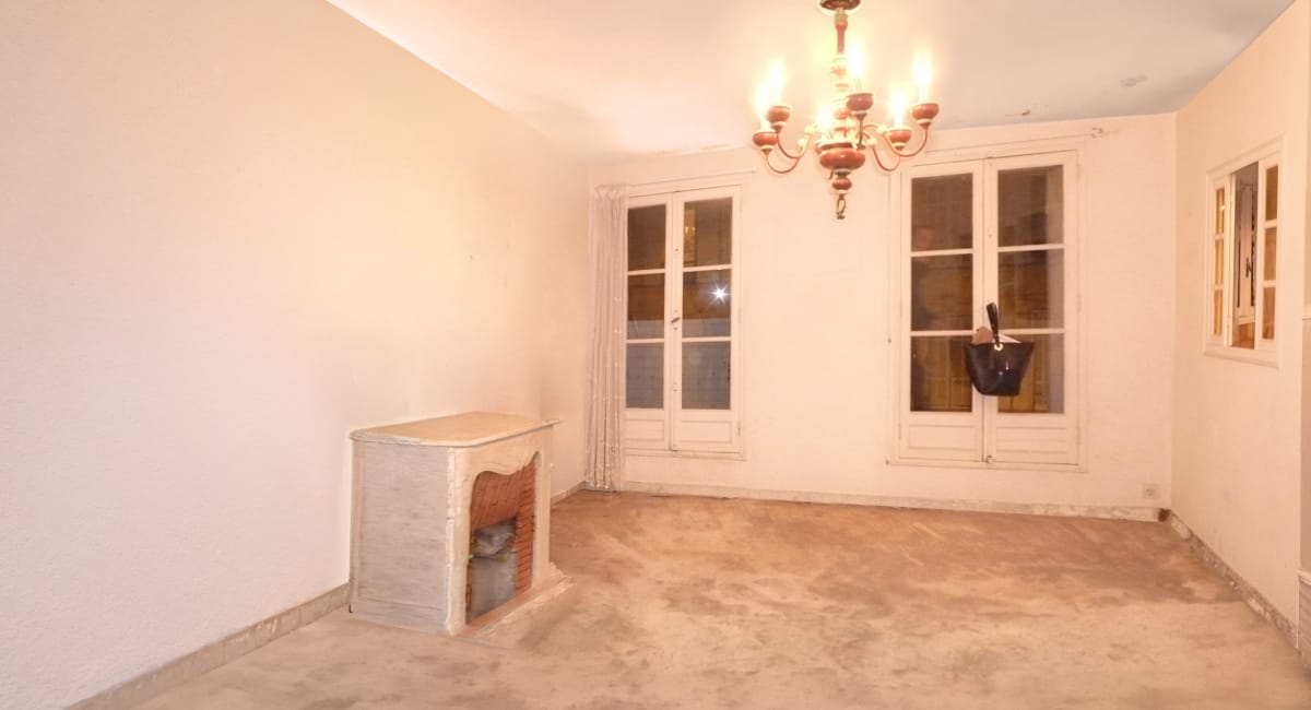 Pièce vide avec cheminée, lustre et grandes fenêtres, à rénover.