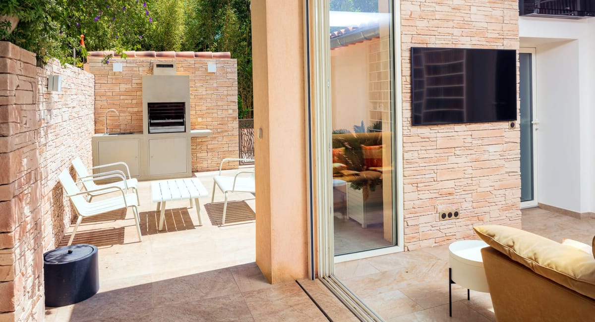 Une terrasse extérieure confortable avec cuisine grill, mobilier blanc et vue sur l'intérieur.