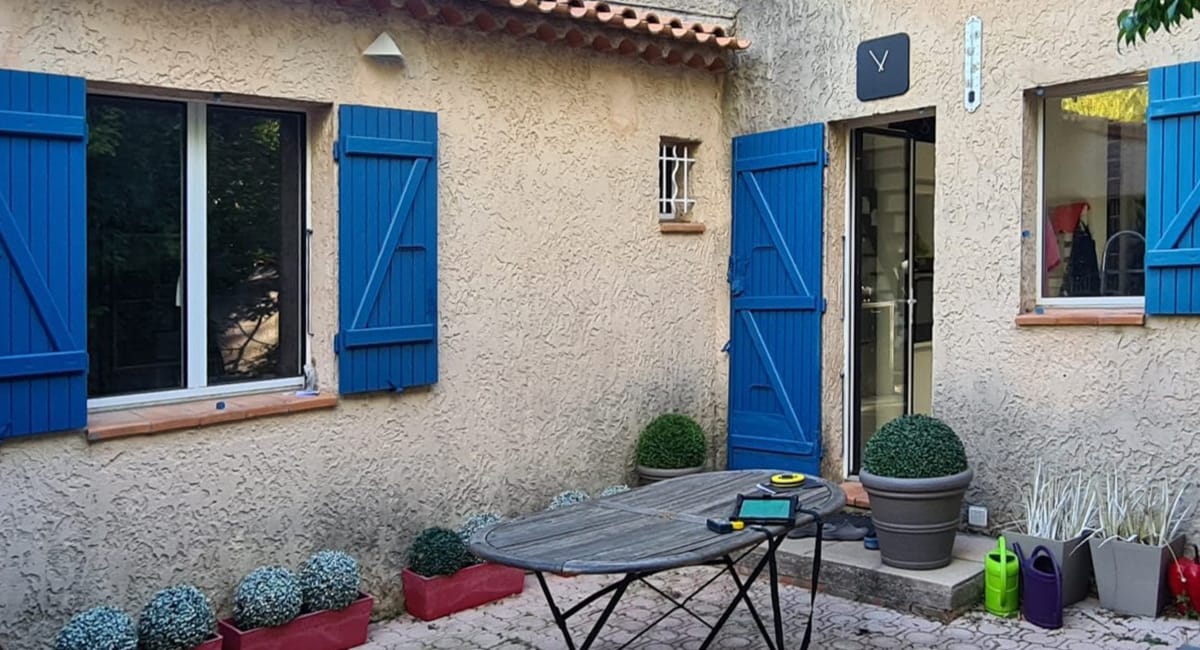 Façade d'une maison avec des volets bleus, des plantes et un petit table.