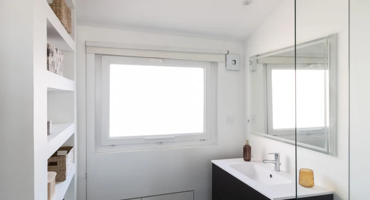 Salle de bain blanche avec deux fenêtres, un miroir et un lavabo.