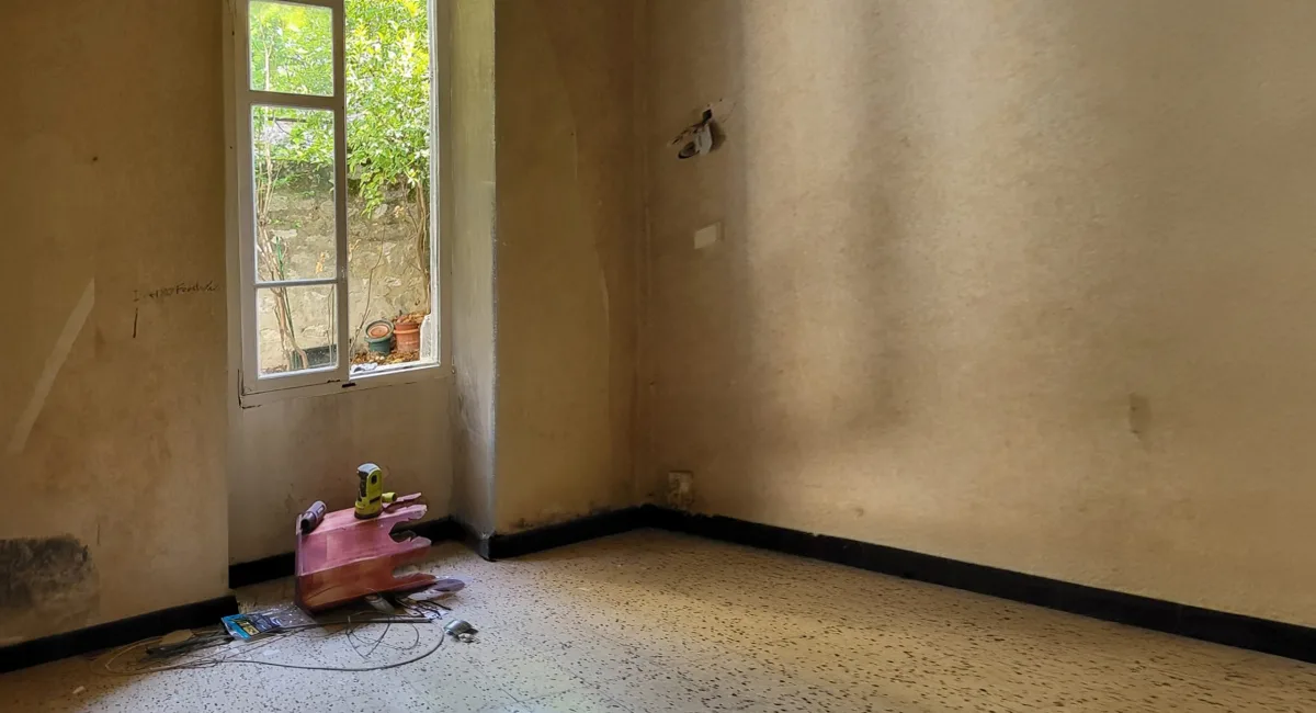 Un coin de pièce délabré avec une fenêtre, un mur beige et un sol carrelé.