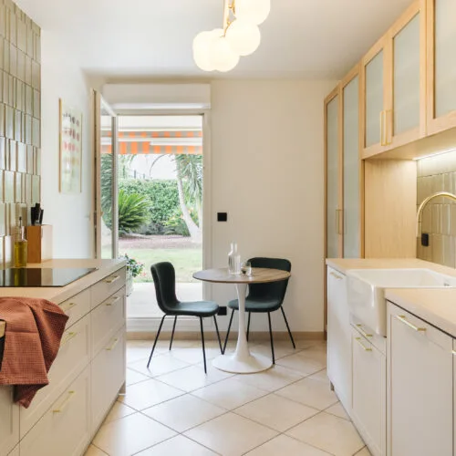 Cuisine moderne et lumineuse avec plan de travail en bois, meubles blancs et un petit coin repas près d'une porte-fenêtre.
